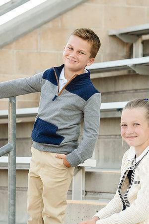 A young boy stands confidently in a gray and navy half-zip pullover, featuring a mock neck and a kanga pocket, with a girl smiling beside him in a casual setting.