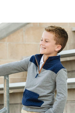 A smiling boy leans against a railing, wearing a gray organic cotton pullover with a navy quilted yoke and half-zip collar.