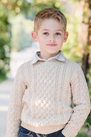 A young boy stands outdoors wearing a beige cable knit sweater and a collared shirt, smiling softly.