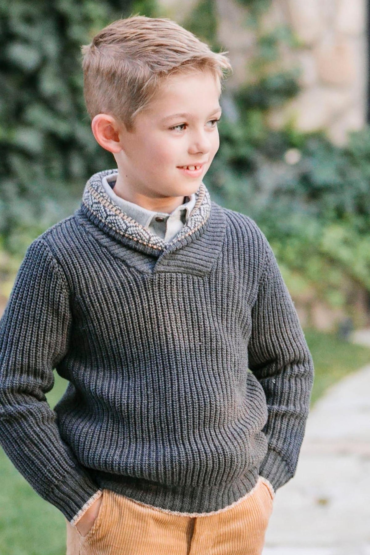 A young boy smiles while wearing a gray shawl collar sweater with a textured design, paired with corduroy pants.