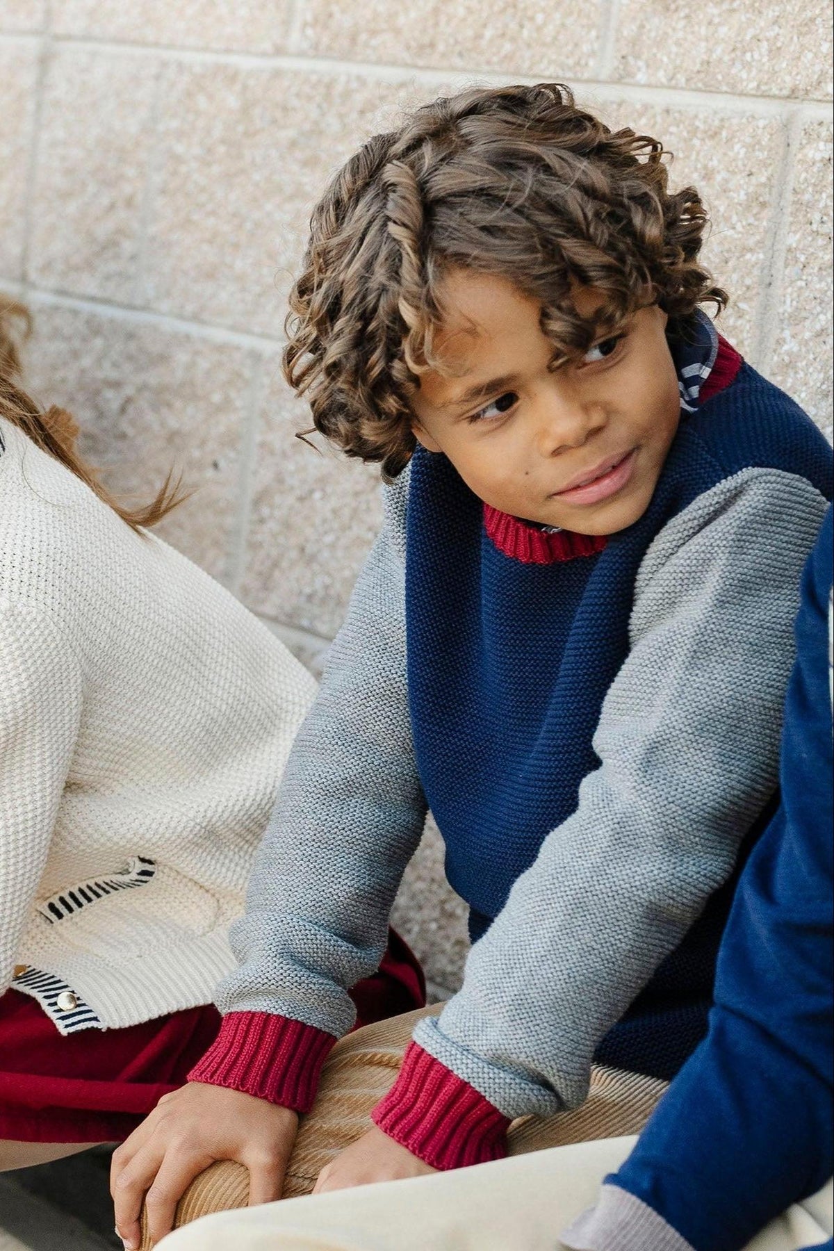 A young boy with curly hair wears a navy and gray crewneck sweater, featuring ribbed red trim, while sitting beside.