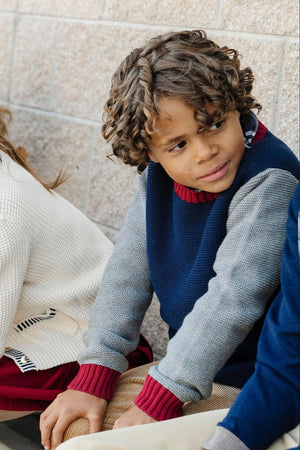 A young boy with curly hair wears a navy and gray crewneck sweater, featuring ribbed red trim, while sitting beside.