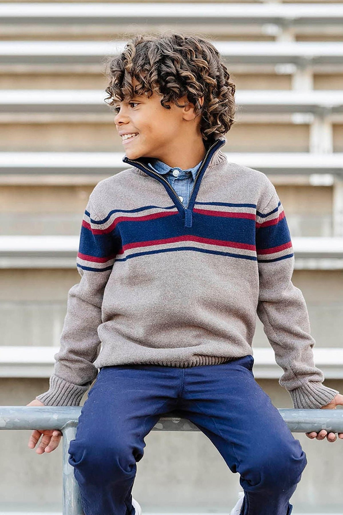 A boy sits on a railing wearing a taupe half-zip sweater with navy and burgundy stripes, paired with navy pants.