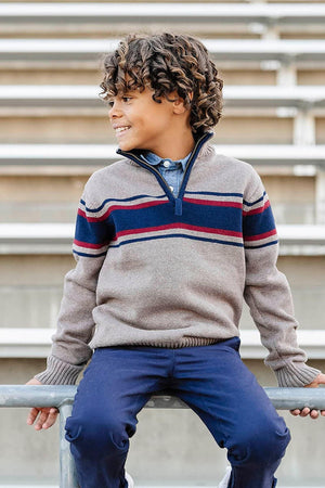 A boy sits on a railing wearing a taupe half-zip sweater with navy and burgundy stripes, paired with navy pants.