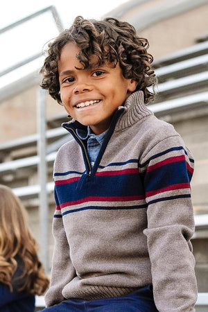A smiling boy with curly hair wears a taupe half-zip sweater featuring blue and red stripes, set against a bleacher.
