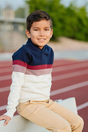 A smiling boy sits on a bench wearing a navy and light gray striped shawl collar sweater and beige pants.