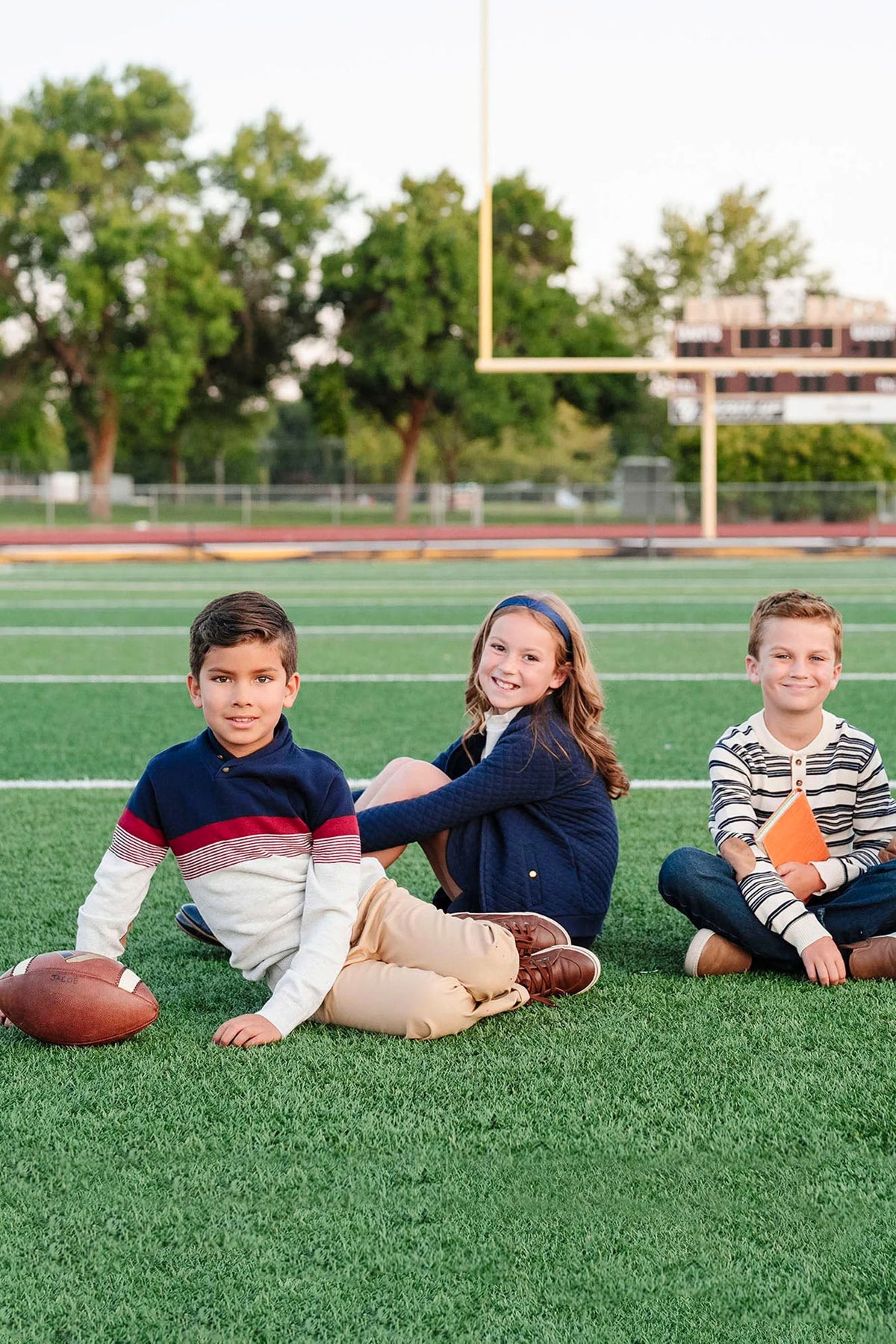 Three children sit on a green turf football field, smiling and wearing casual clothing.