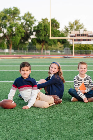 Three children sit on a green turf football field, smiling and wearing casual clothing.