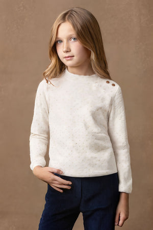 A girl wearing a soft white mock neck knit top with gold buttons at the shoulder stands against a textured brown background.