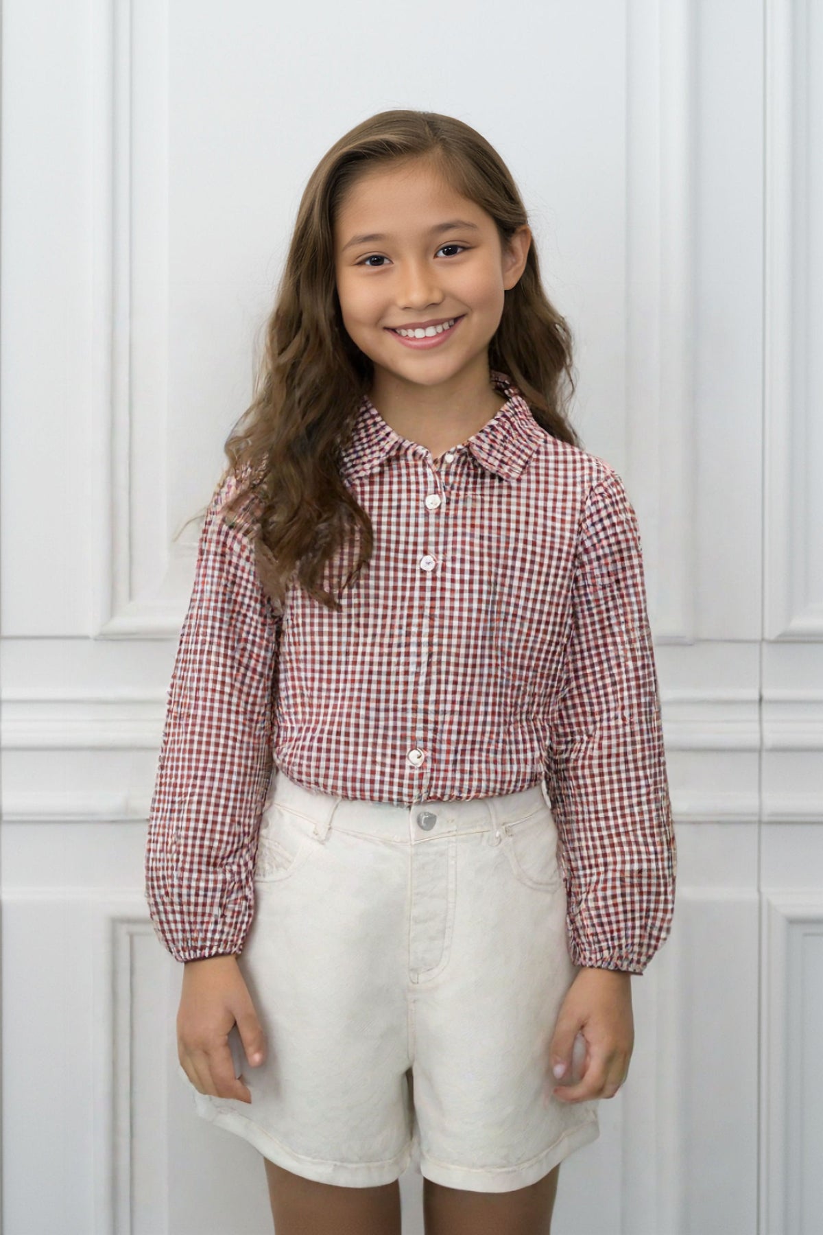 A smiling girl wearing a red and white gingham button-up shirt and white shorts stands against a light backdrop.