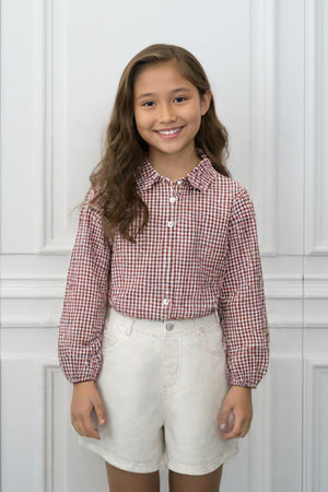 A smiling girl wearing a red and white gingham button-up shirt and white shorts stands against a light backdrop.
