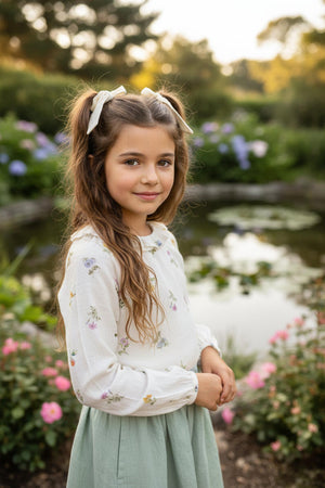 A girl with long hair stands by a pond, wearing an ivory floral top and a mint green skirt, surrounded.