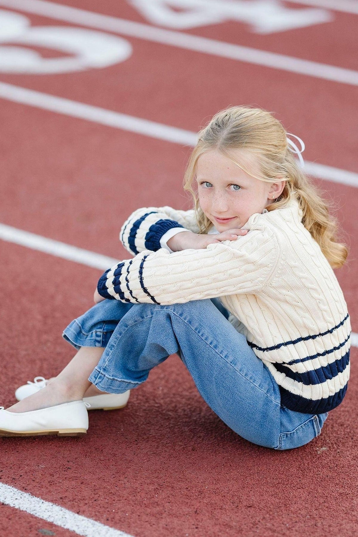 A girl sits on a red track wearing a cozy ivory roll neck sweater with navy stripes and blue jeans.