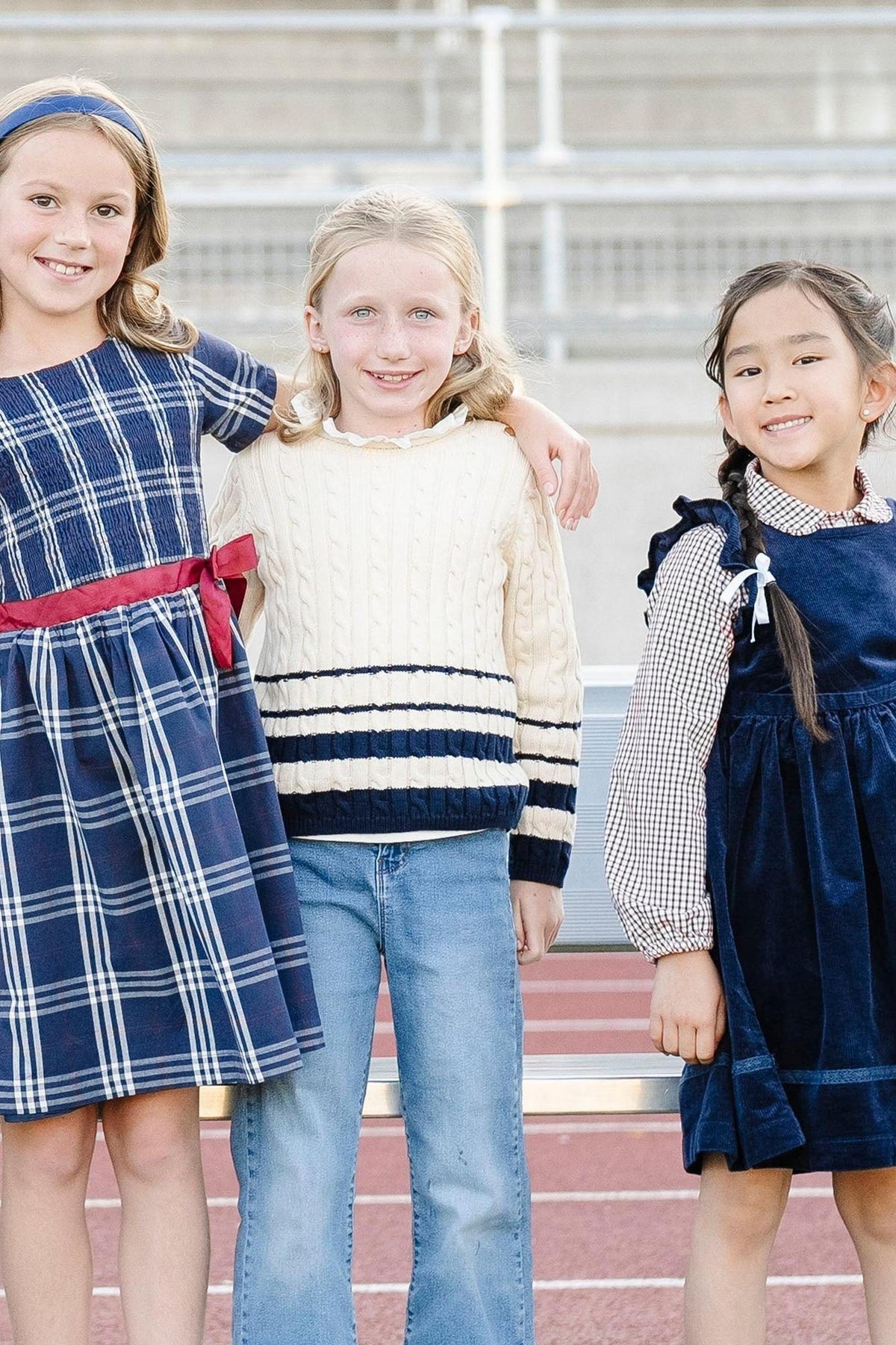 Three girls stand together, wearing stylish outfits: a plaid dress, a cream cable-knit sweater, and a navy corduroy dress.