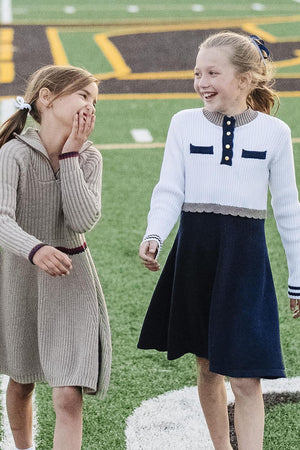 Two young girls playfully walk on a football field, wearing stylish sweater dresses. One dress is beige with a mock neckline, while the other features a white and navy design.