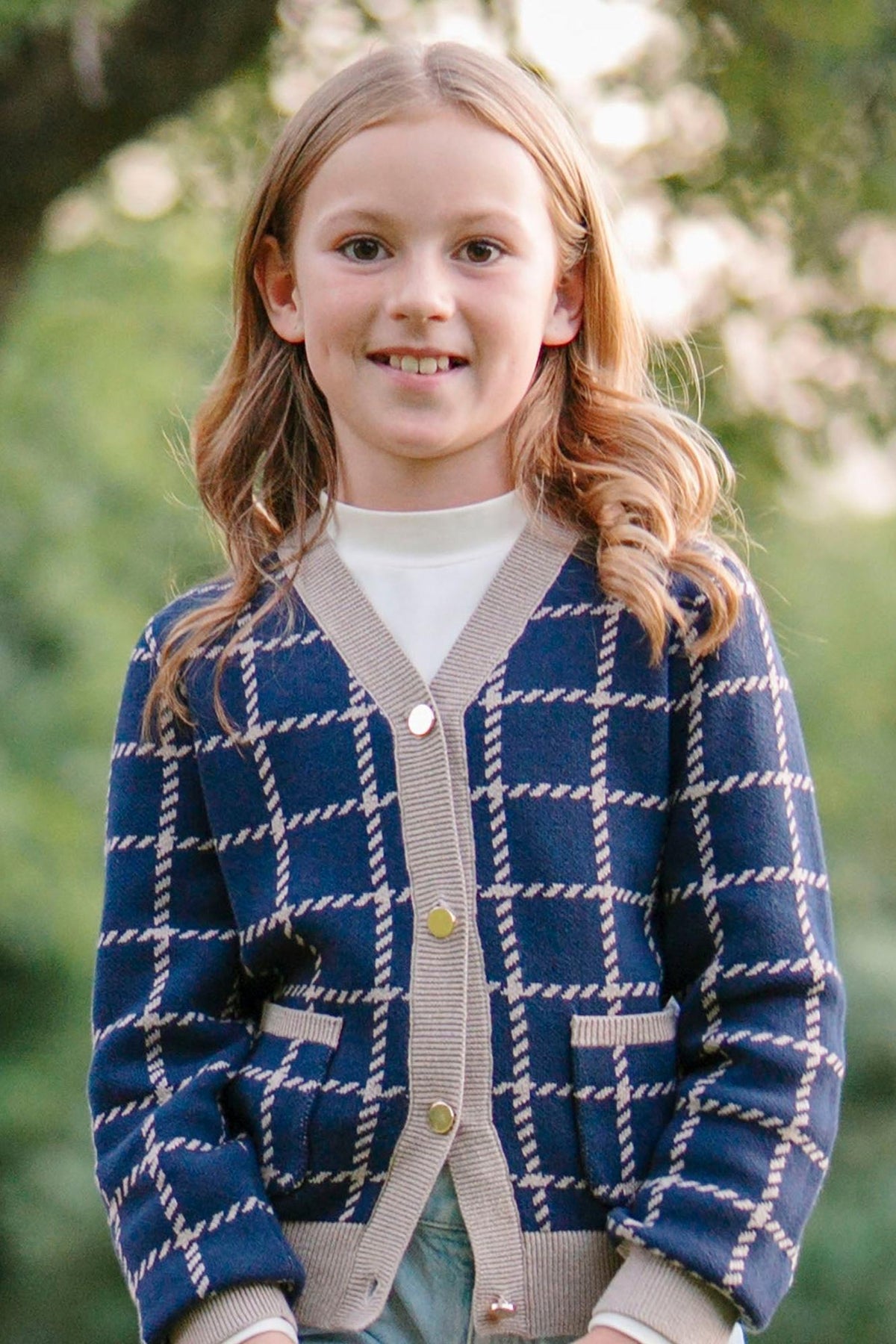 A young girl smiles while wearing a navy and tan plaid cardigan with front pockets and gold buttons, outdoors.