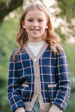 A young girl smiles while wearing a navy and tan plaid cardigan with front pockets and gold buttons, outdoors.