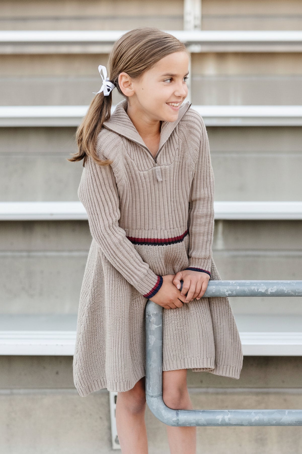 A girl smiles while leaning against a railing, wearing a taupe heather rib knit half zip sweater dress with a contrasting stripe at the waist, showcasing its organic cotton material.