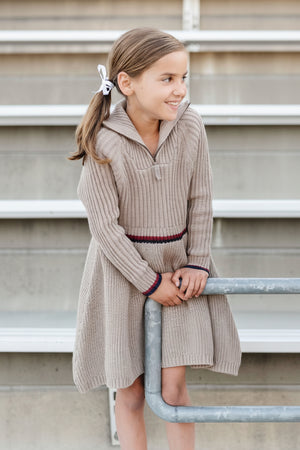 A girl smiles while leaning against a railing, wearing a taupe heather rib knit half zip sweater dress with a contrasting stripe at the waist, showcasing its organic cotton material.