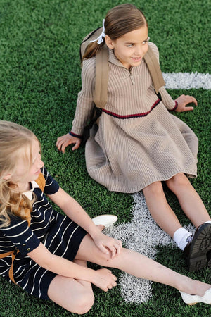 Two young girls sit on green grass, one wearing a taupe heather rib knit sweater dress with a half zip and contrast stripe, while the other wears a navy striped.