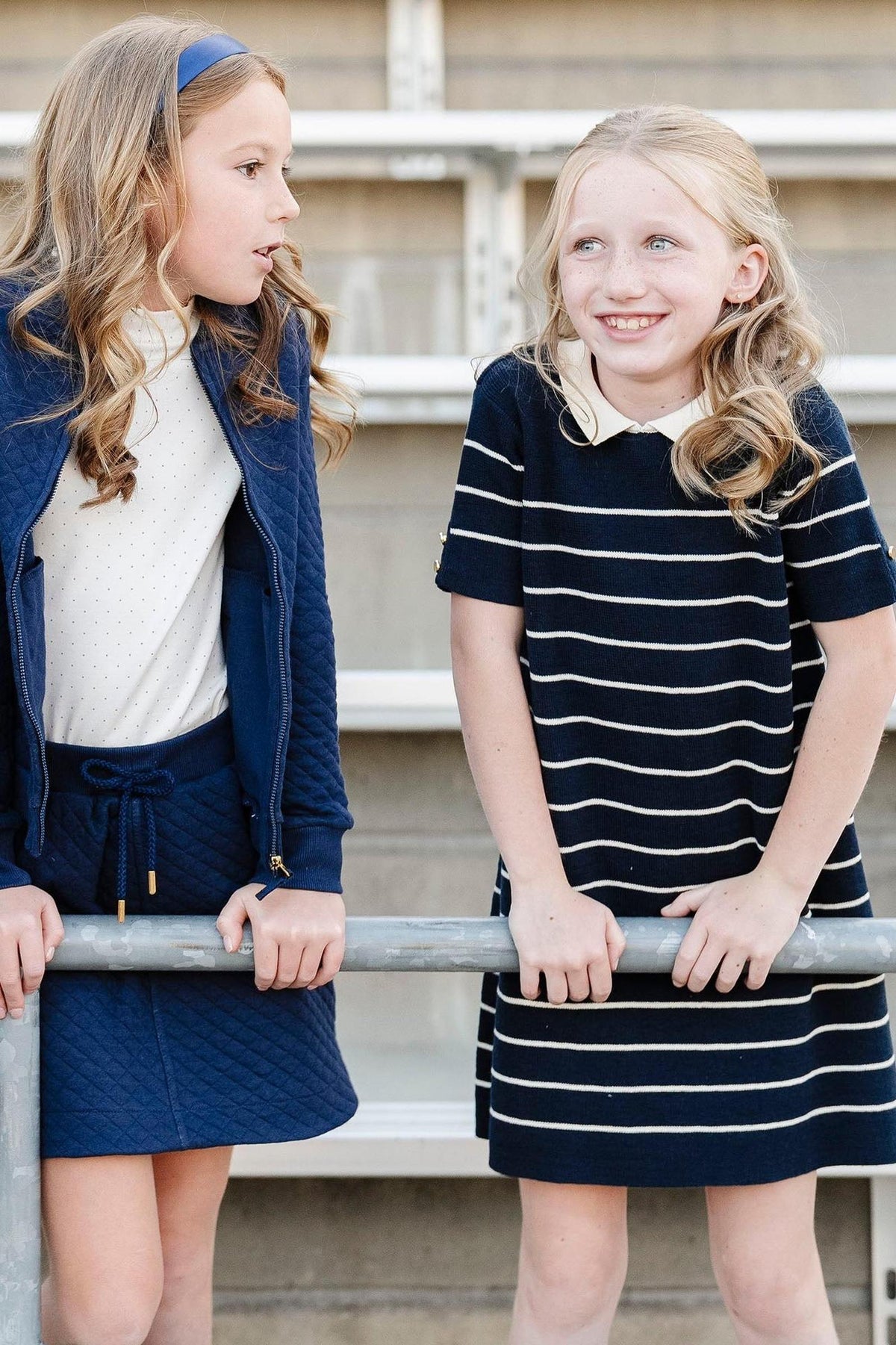Two young girls wearing navy outfits, one in a striped dress with a collar, smiling and chatting outdoors.