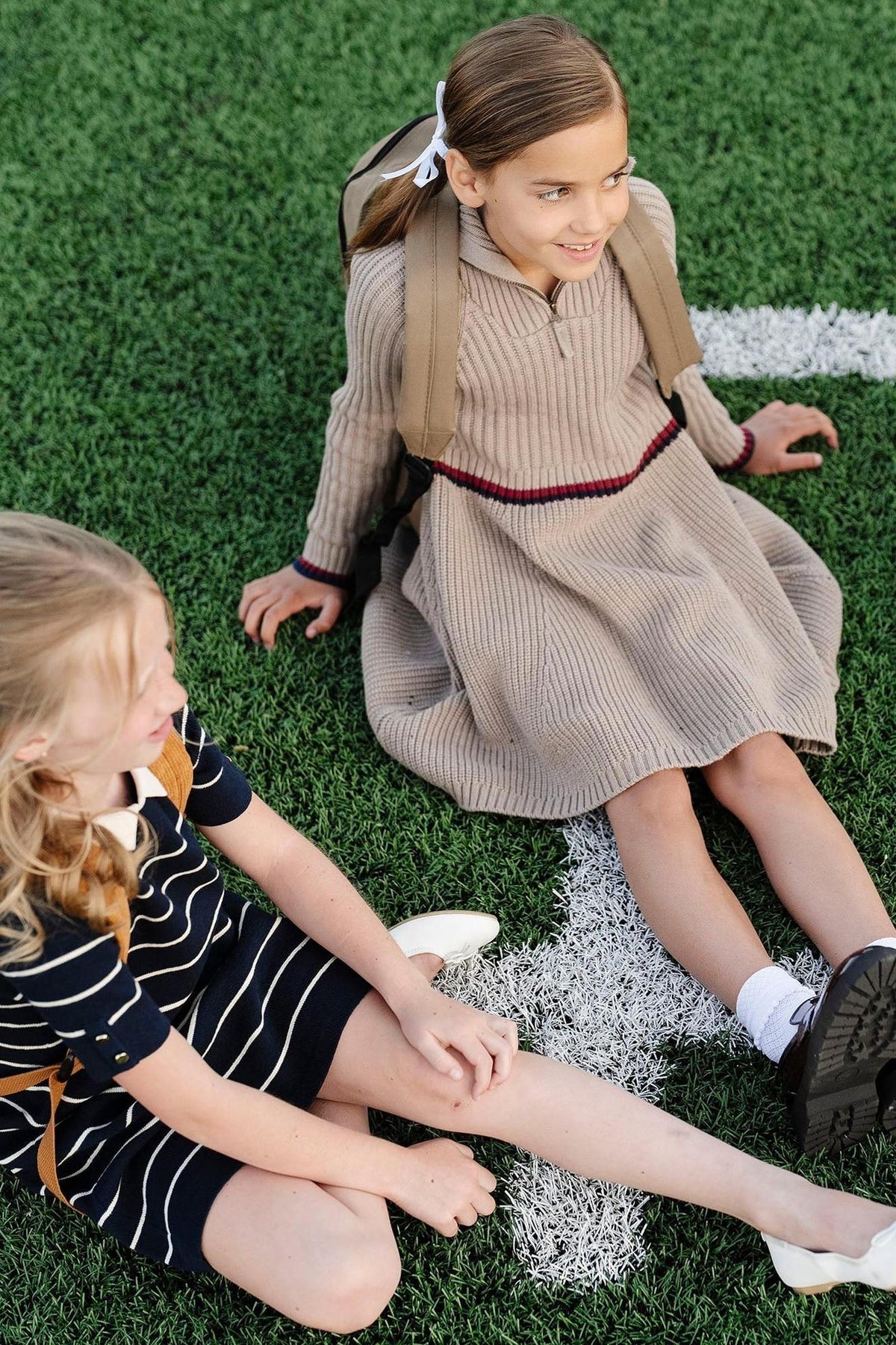 Two girls sitting on grass; one wears a beige ribbed dress, the other a navy striped dress with backpacks.