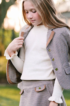A young girl wears an ivory organic cotton sweater with ruffled edges, paired with a houndstooth blazer and skirt.