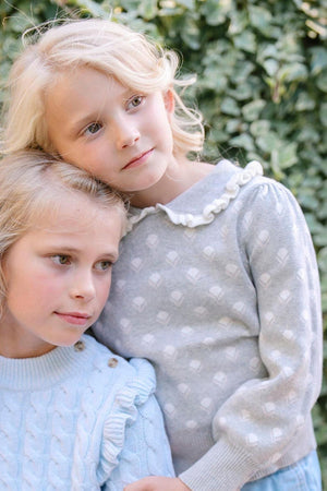 Two young girls are posing together outdoors, wearing soft sweaters; one in light gray with floral patterns and the other.