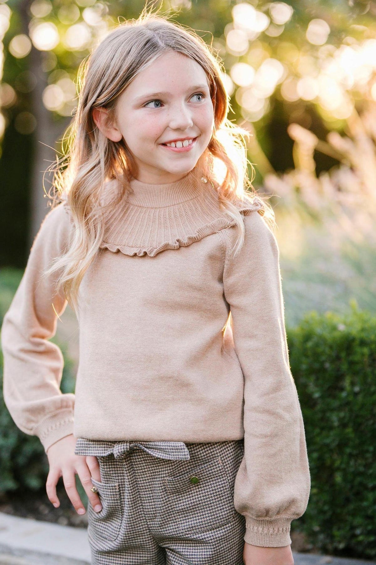 A young girl smiles while wearing a camel heather mock neck sweater with ruffle detailing. The soft, organic cotton sweater is paired with stylish pants, set against a sunlit outdoor.