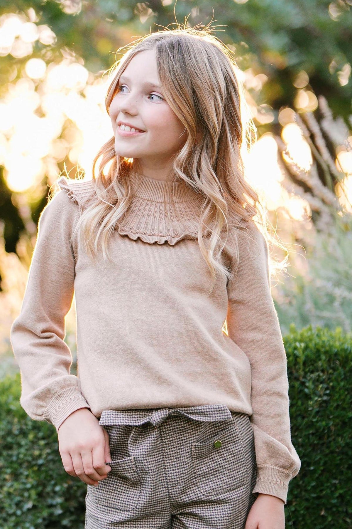 A girl smiles while wearing a soft, camel heather organic cotton mock neck sweater with ruffle details. The background features greenery and warm sunlight, enhancing the cozy atmosphere.