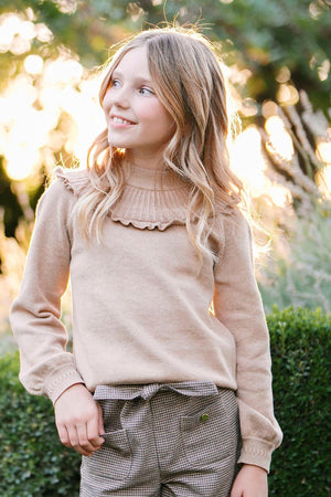 A girl smiles while wearing a soft, camel heather organic cotton mock neck sweater with ruffle details. The background features greenery and warm sunlight, enhancing the cozy atmosphere.