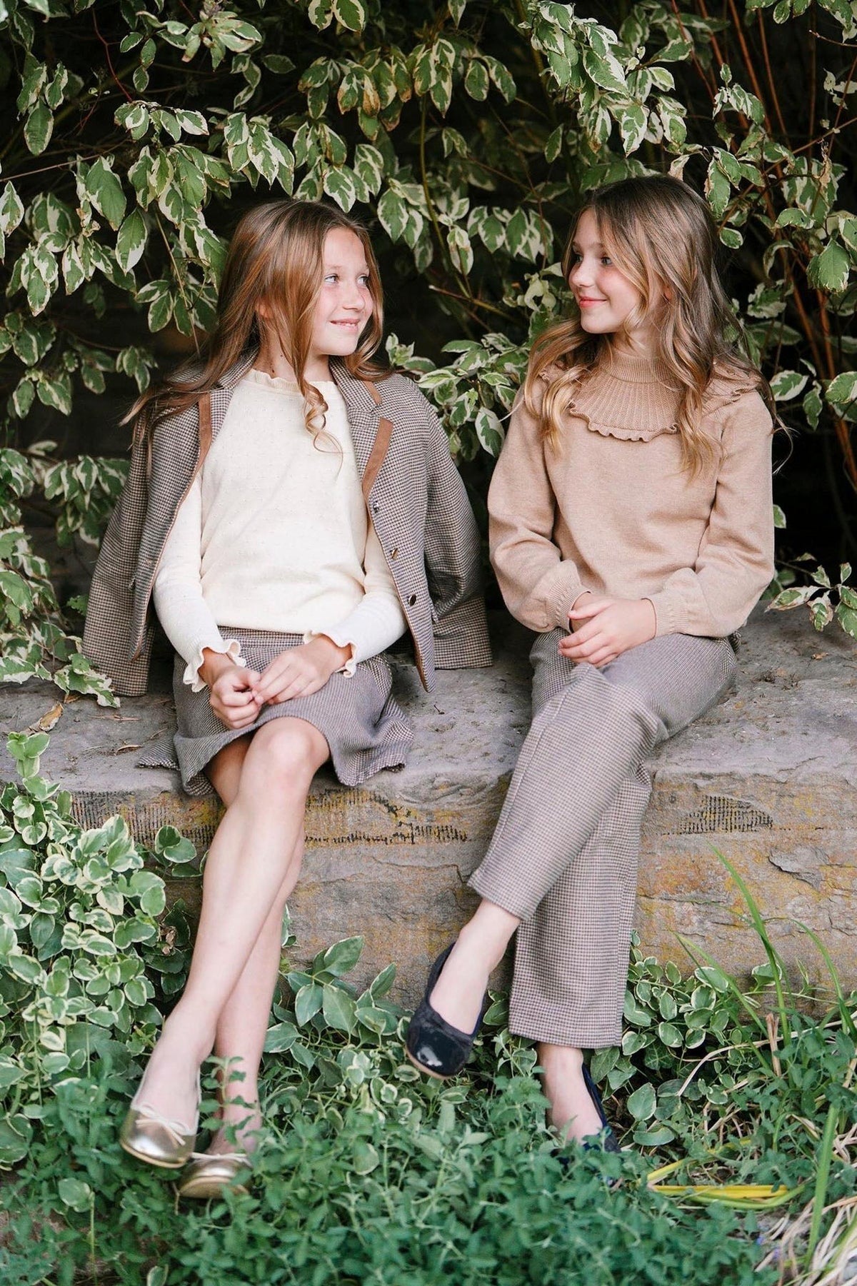 Two girls sit on a stone ledge surrounded by greenery, wearing stylish outfits. One girl is in a cream sweater and skirt, while the other wears a camel ribbed sweater.