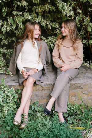 Two girls sit on a stone ledge surrounded by greenery, wearing stylish outfits. One girl is in a cream sweater and skirt, while the other wears a camel ribbed sweater.