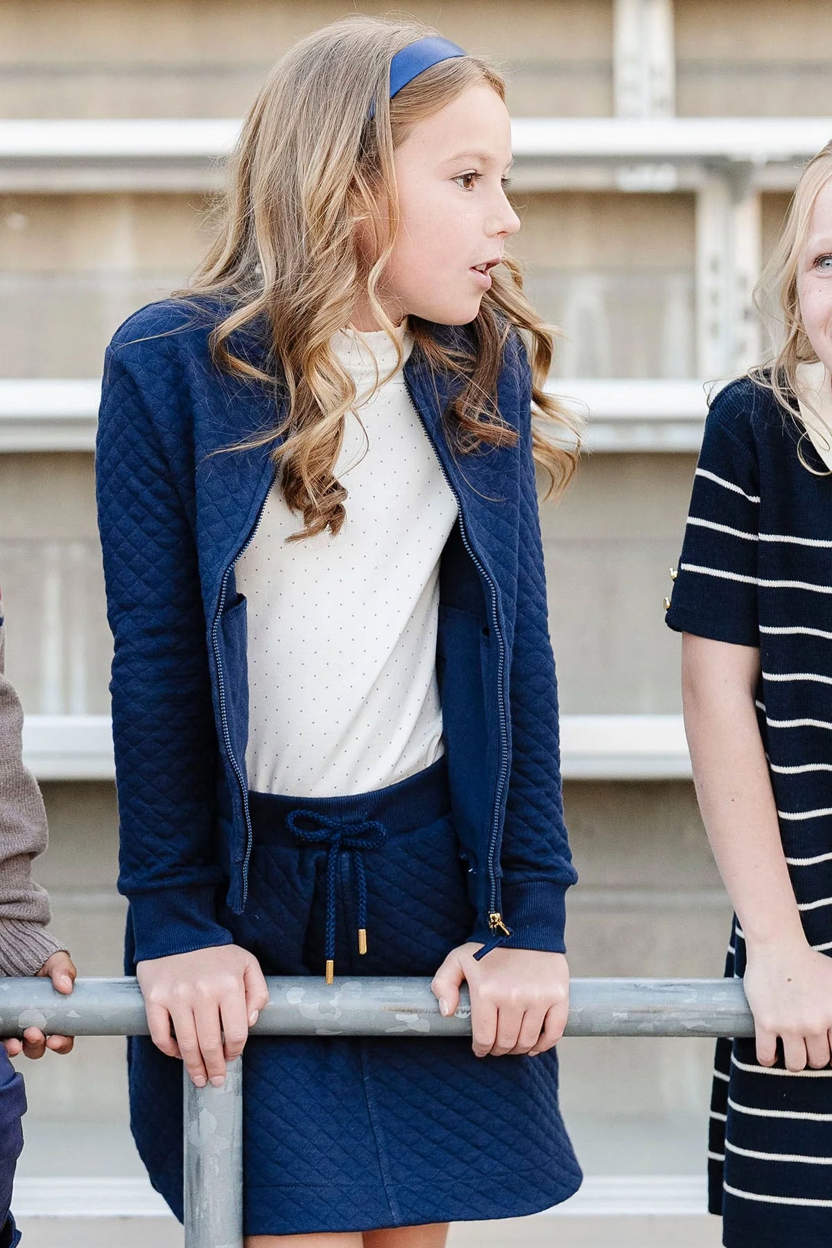 A girl in a navy quilted jacket and matching skort leans on a railing, surrounded by friends in casual outfits.