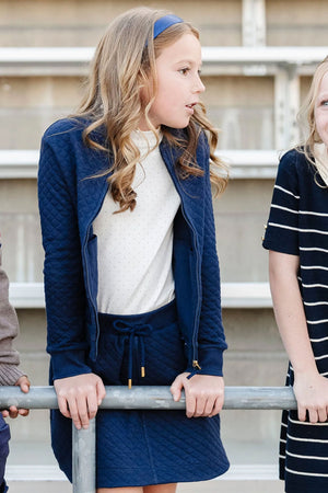 A girl in a navy quilted jacket and matching skort leans on a railing, surrounded by friends in casual outfits.
