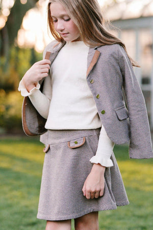 A girl wears a houndstooth skirt and matching blazer, styled with a cream sweater, in a garden setting.
