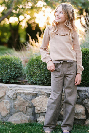 A young girl stands outside in a camel-colored sweater and houndstooth wide-leg pants, smiling amidst greenery and soft sunlight.