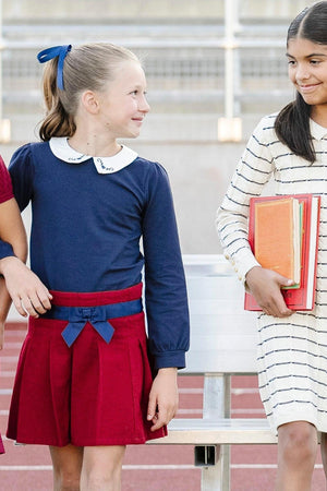 A girl wearing a navy long-sleeve top and a red pleated skirt with a blue bow stands next to another.
