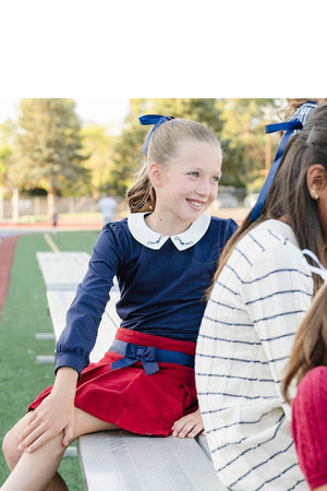 A young girl smiles while sitting on bleachers, wearing a navy top and a red pleated skirt with a blue.
