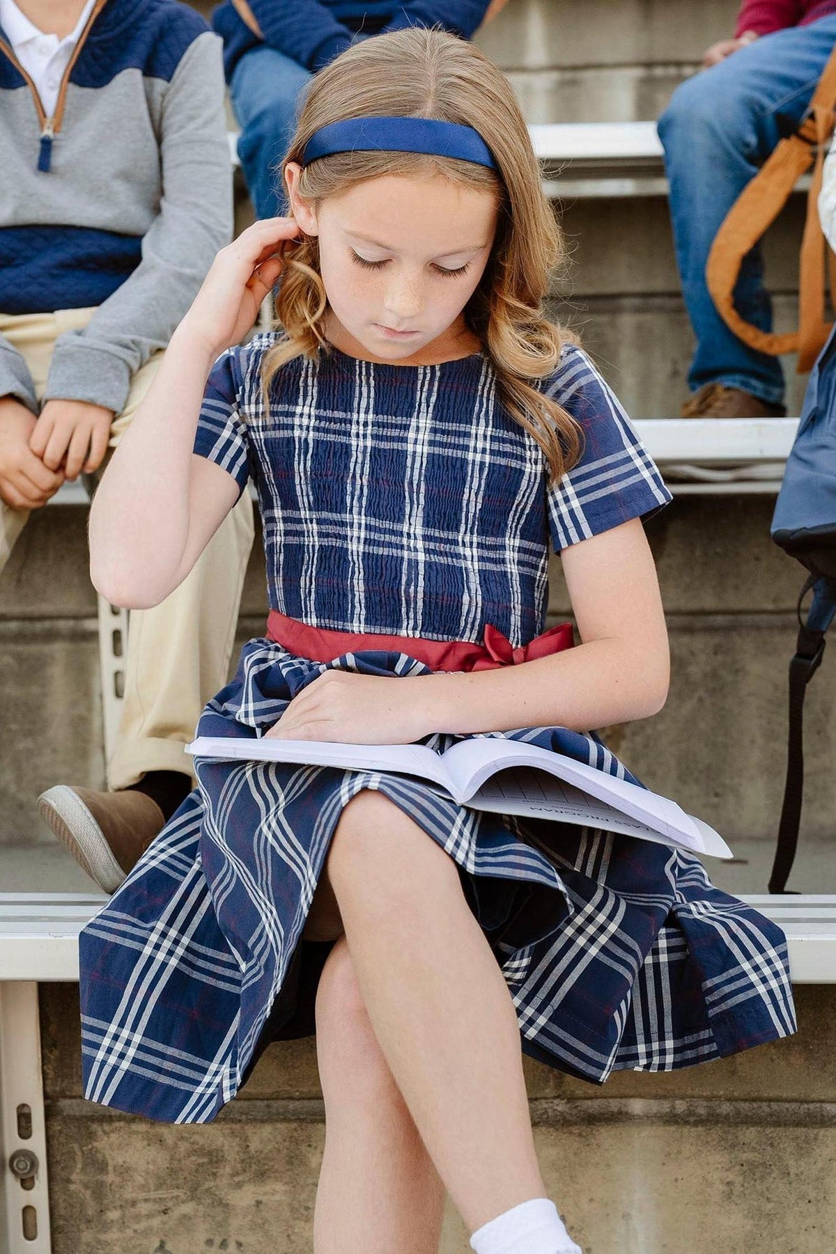 A girl sits on bleachers wearing a navy plaid dress with short sleeves and a red belt, reading a notebook.