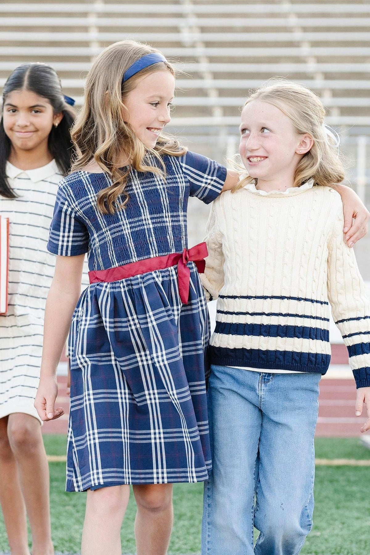 Two girls are smiling while wearing a navy plaid dress with a red belt and a cream-colored cable-knit sweater.