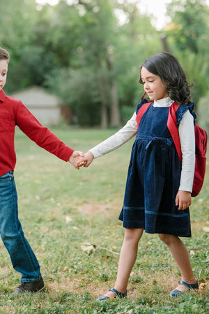 A girl in a navy jumper dress with flutter sleeves holds hands with a boy in a red shirt, outdoors.