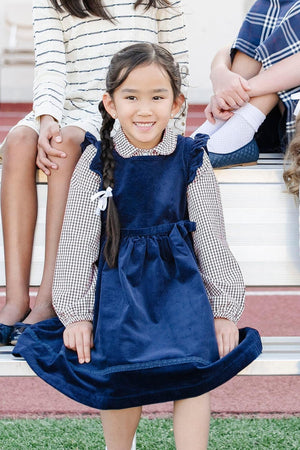 A young girl smiles while wearing a navy blue corduroy jumper dress with flutter sleeves, paired with a checkered shirt.