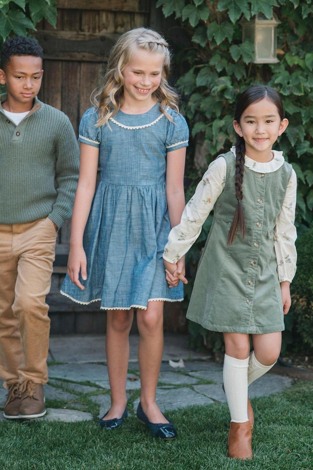 Three children are walking hand in hand, wearing stylish dresses and a sweater, against a lush green backdrop.