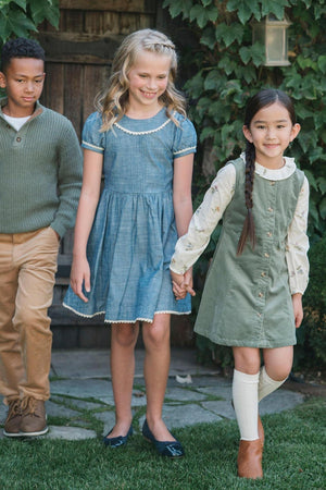 Three children are walking hand in hand, wearing stylish dresses and a sweater, against a lush green backdrop.