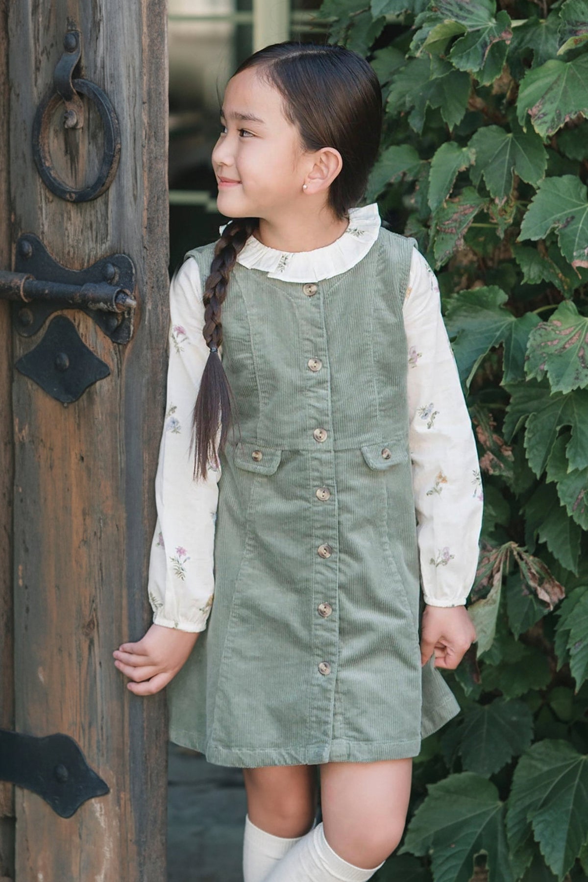 A girl smiles while wearing a sage green corduroy jumper dress over a floral long-sleeve shirt, standing beside a wooden.