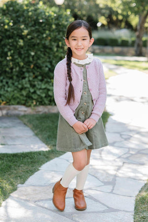 A young girl stands on a stone pathway, wearing a sage green corduroy jumper dress, paired with a light pink.