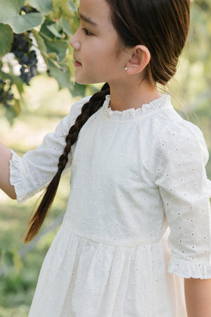 A child in an ivory, organic cotton eyelet dress with ruffle details stands outdoors near grapevines.