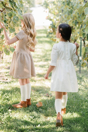 Two children wearing dresses and boots walk through a vineyard, surrounded by lush green leaves and grass.
