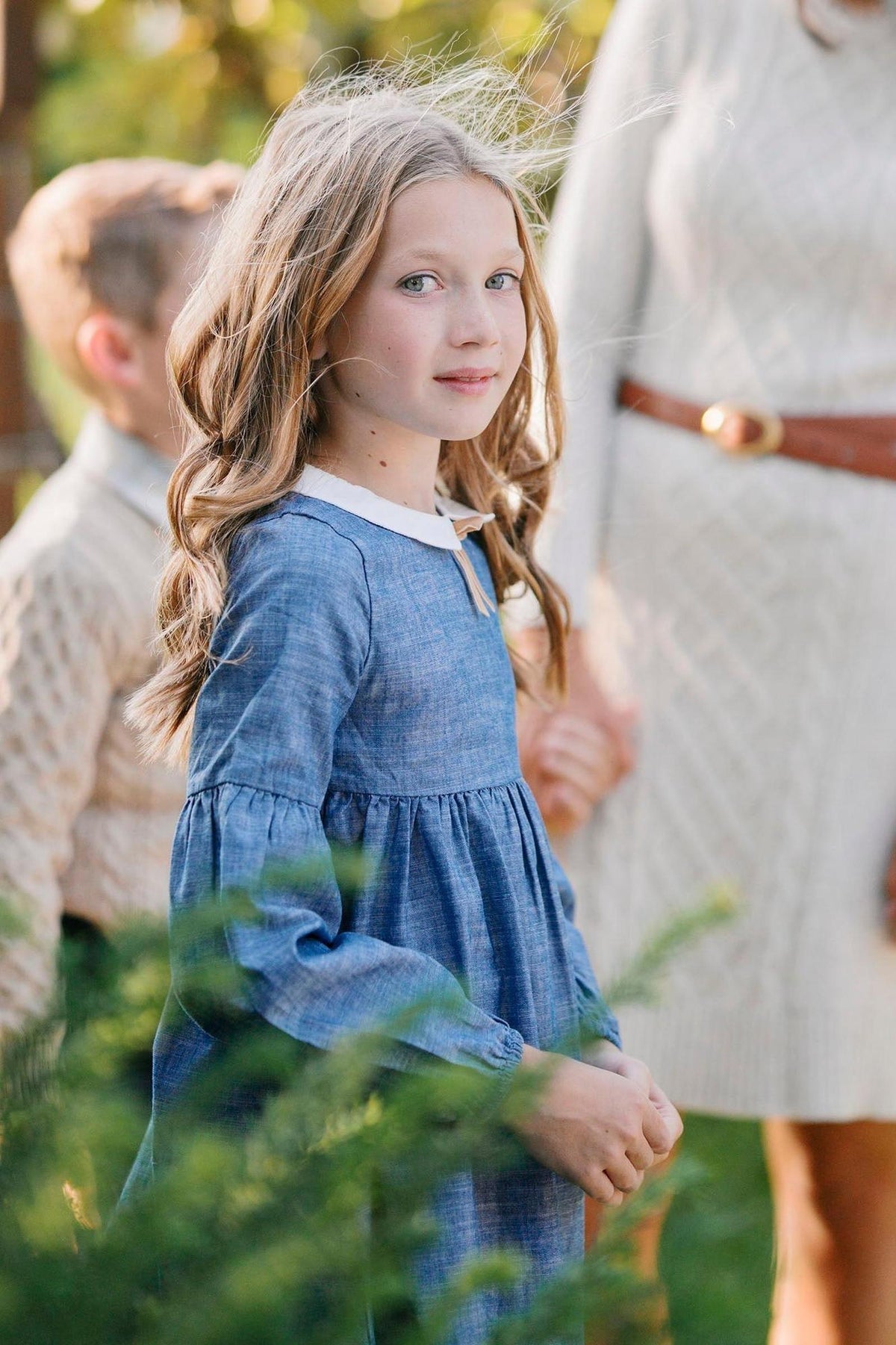 A young girl with long, wavy hair wears a chambray dress featuring a Peter Pan collar and puffed sleeves, standing amidst greenery.